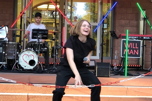 Eine junge Frau mit gebeugten Knien hält ein Mikrofon in der Hand. Im Hintergrund spielt ein junger Mann Schlagzeug und es sind rote, blaue und grüne LED-Lichter zu sehen.